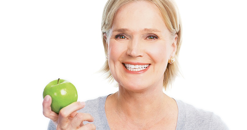 An Old Lady Holding A Green Apple On Right Hand Looking At Camera And Showing Her Teeth