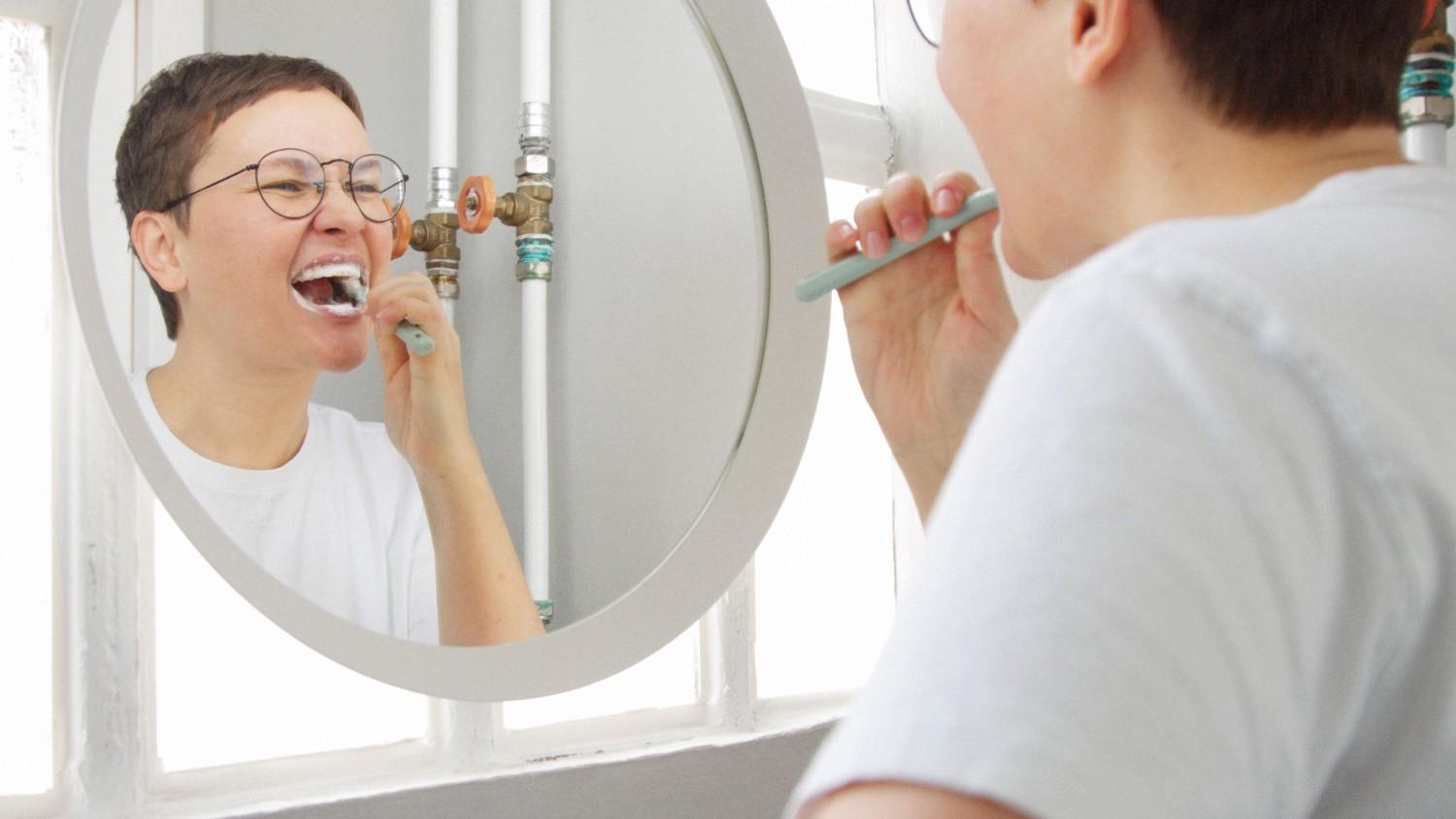 person brushing teeth in mirror for brighter smile